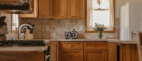 Una cocina con armarios de madera, salpicadero de azulejos, una ventana con cortina y suelo de ladrillo.