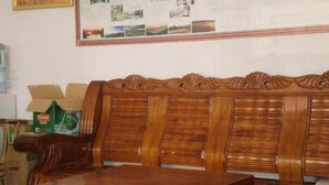Un banco de madera con respaldo alto, una mesa al frente y un tablón de anuncios con fotos y texto en la pared de fondo.
