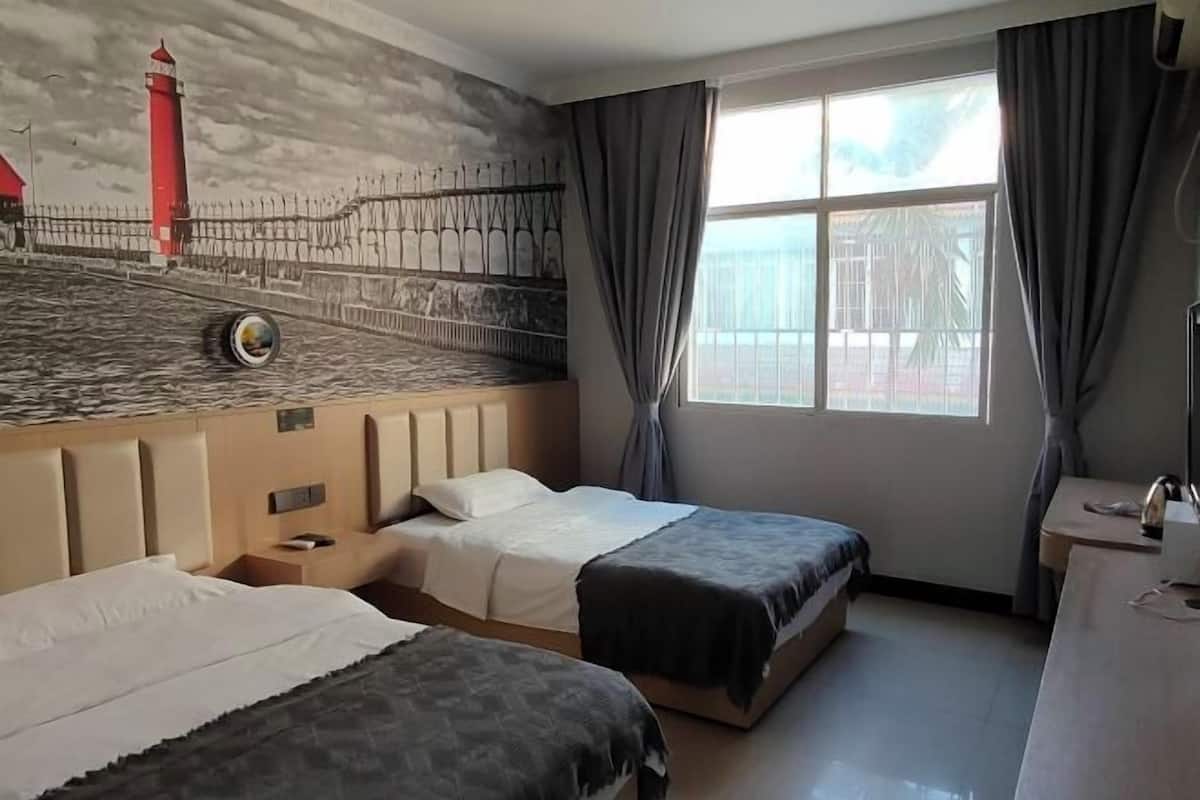 Une chambre d’hôtel avec deux lits, une grande fenêtre avec des rideaux, une fresque murale représentant un phare et un bureau avec une lampe.