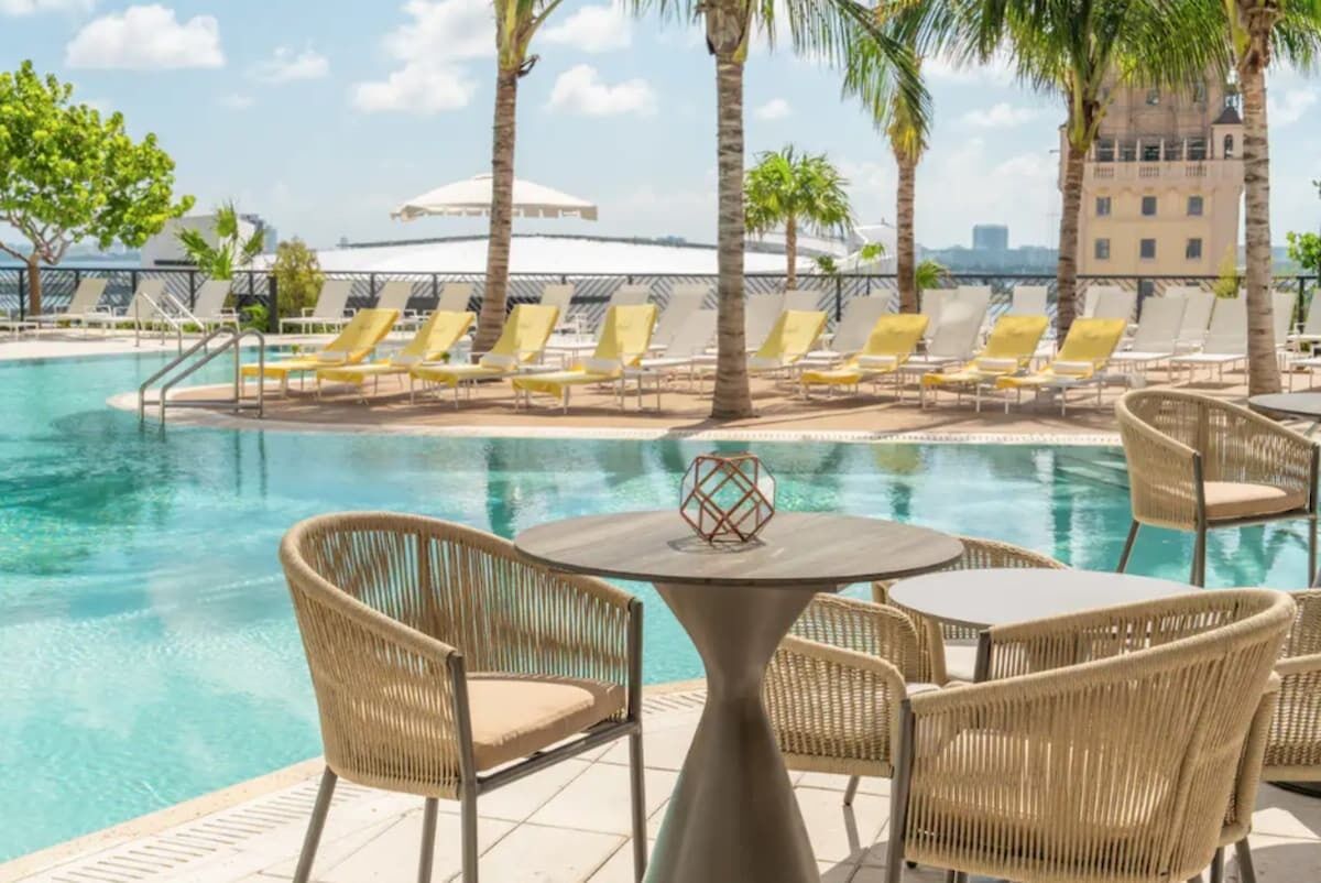 Un espace aménagé au bord de la piscine, avec des chaises longues et une table, entouré de palmiers et offrant une vue dégagée sur les gratte-ciel de la ville.