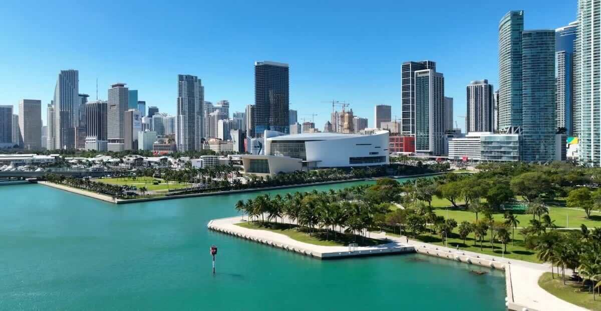 A city skyline with modern high-rise buildings, a marina with boats, and a park with palm trees.