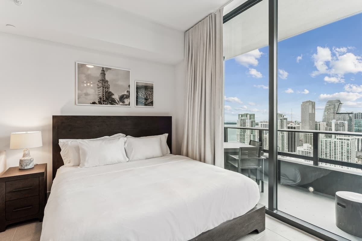 A modern hotel room with a large bed, a nightstand with a lamp, and a city view through large windows.