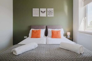A double bed with white and orange pillows, a patterned bedspread, and a view of the outside through a window. - Large 3 Bed Home With 2 Living Areas & Sofa Bed (Northampton)