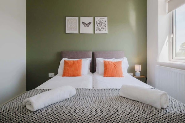 A double bed with white and orange pillows, a patterned bedspread, and a view of the outside through a window.
