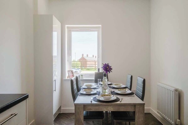 A dining area with a table set for four, a window with a view, and a radiator on the wall. - Large 3 Bed Home With 2 Living Areas & Sofa Bed (Northampton)