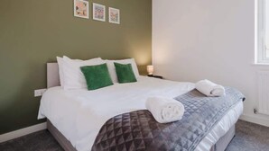 A neatly made bed with white and green pillows, a grey quilt, and a rolled towel on the bed. - Spacious 3 Bed Home With 2 Living Areas & Sofa Bed (Northampton)