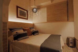 Une chambre à coucher avec un lit, des oreillers, une étagère fixée au mur, un tableau encadré, un luminaire suspendu et une poubelle.