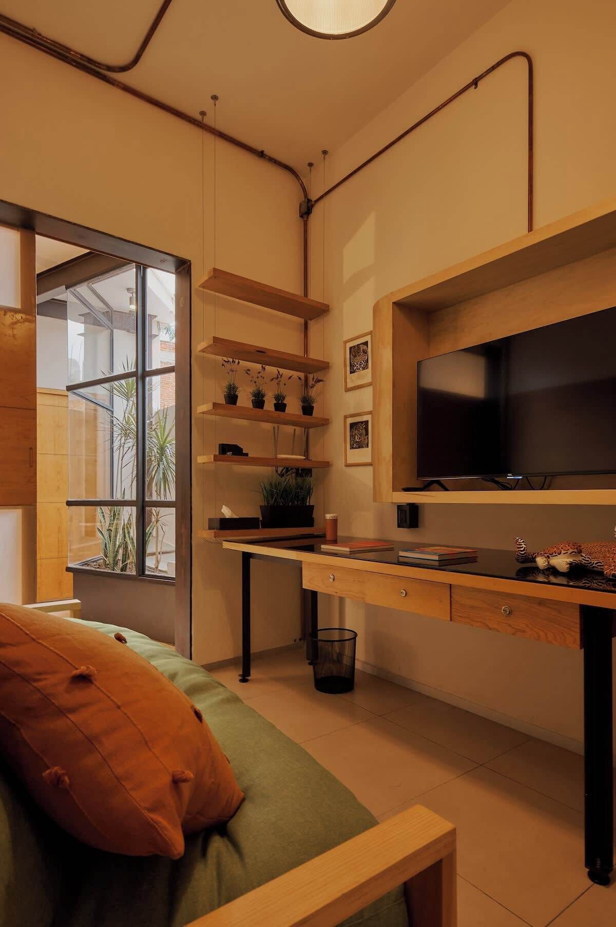 A compact living space with a bed, a desk, and a TV. There are shelves with plants and a window providing natural light.