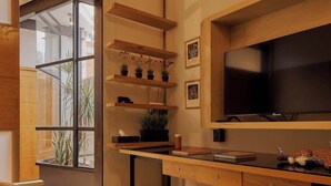 A compact living space with a bed, a desk, and a TV. There are shelves with plants and a window providing natural light.