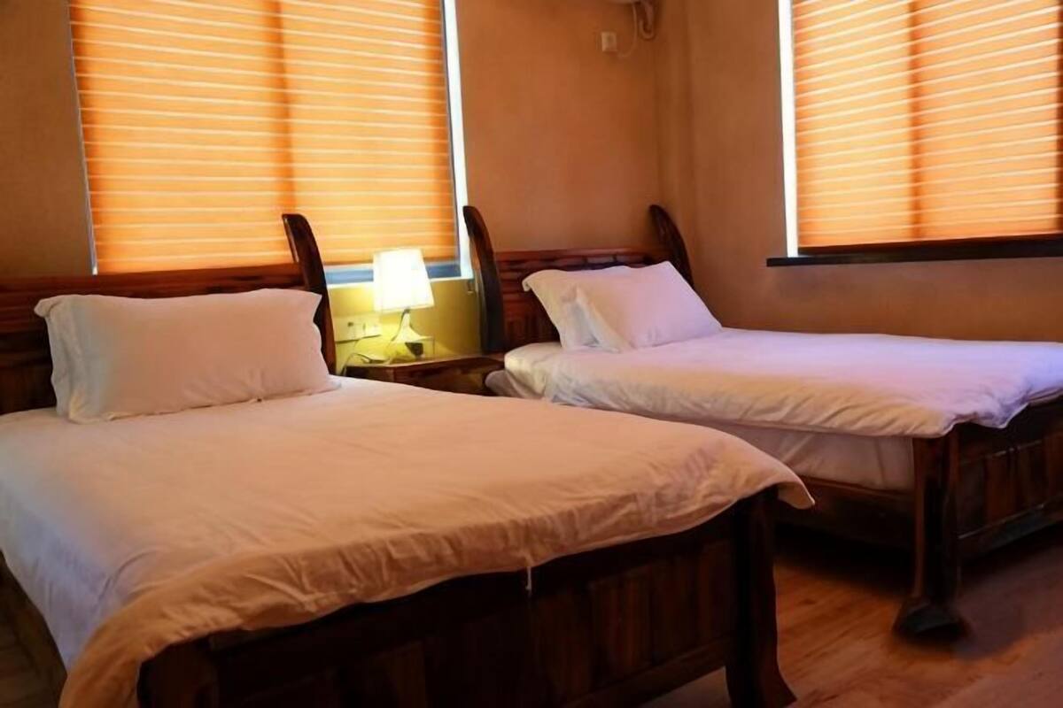 Two single beds with wooden frames and white bedding, a bedside table with a lamp, and windows with blinds.