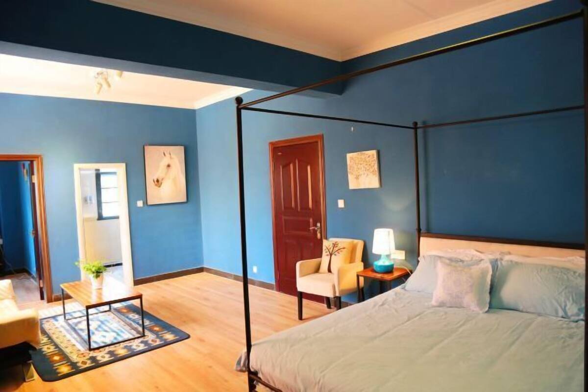 A bedroom with a four-poster bed, a chair, a nightstand with a lamp, and a door.