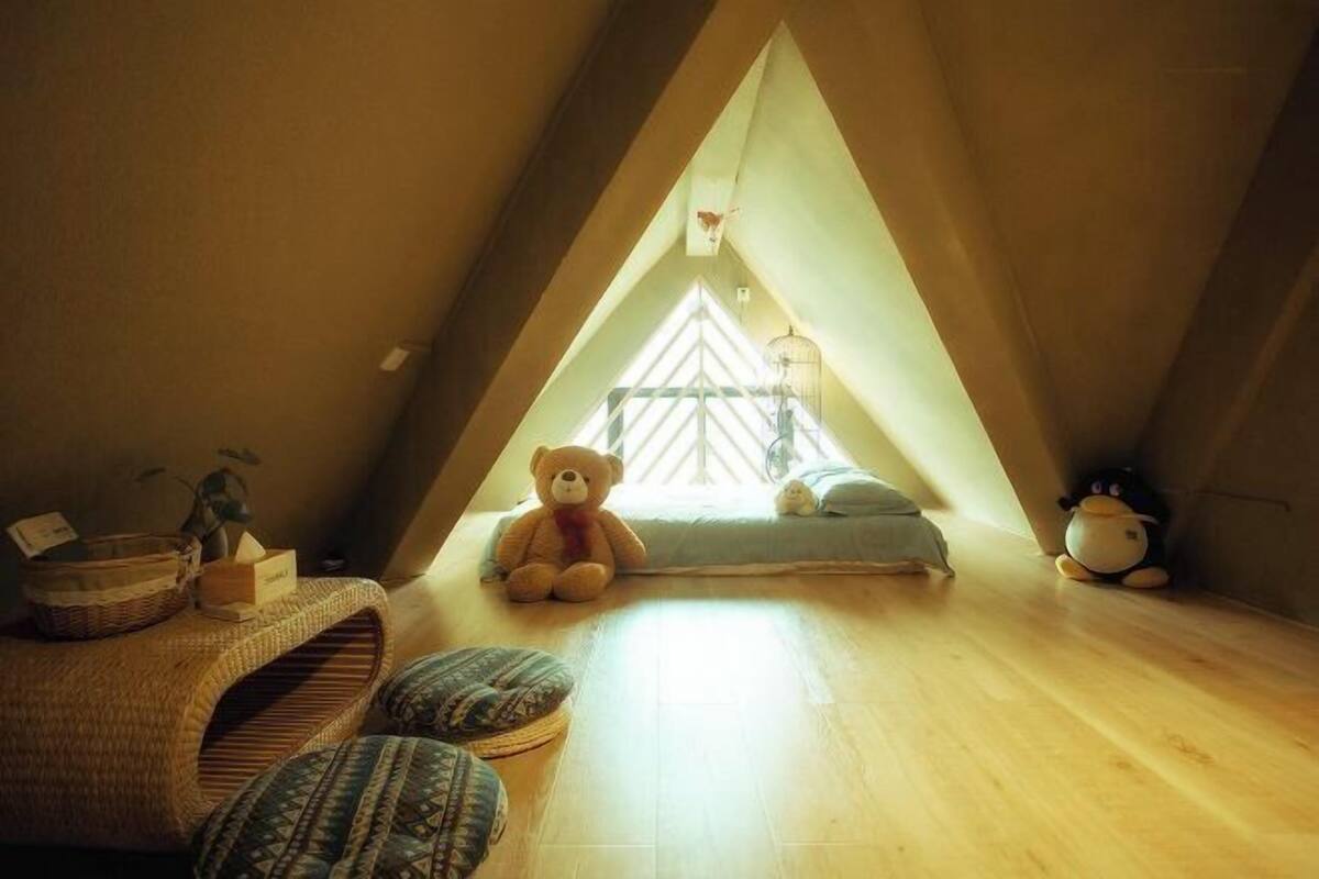 A cozy attic bedroom with a sloped ceiling, a large window, a teddy bear, a plush penguin, and a small bed.