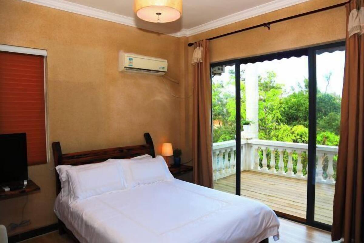 A bedroom with a bed, an air conditioning unit, a TV, and a balcony with a view of greenery.