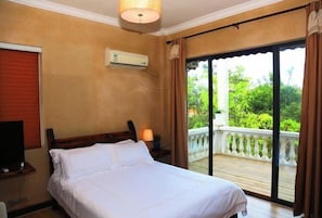 A bedroom with a bed, an air conditioning unit, a TV, and a balcony with a view of greenery.