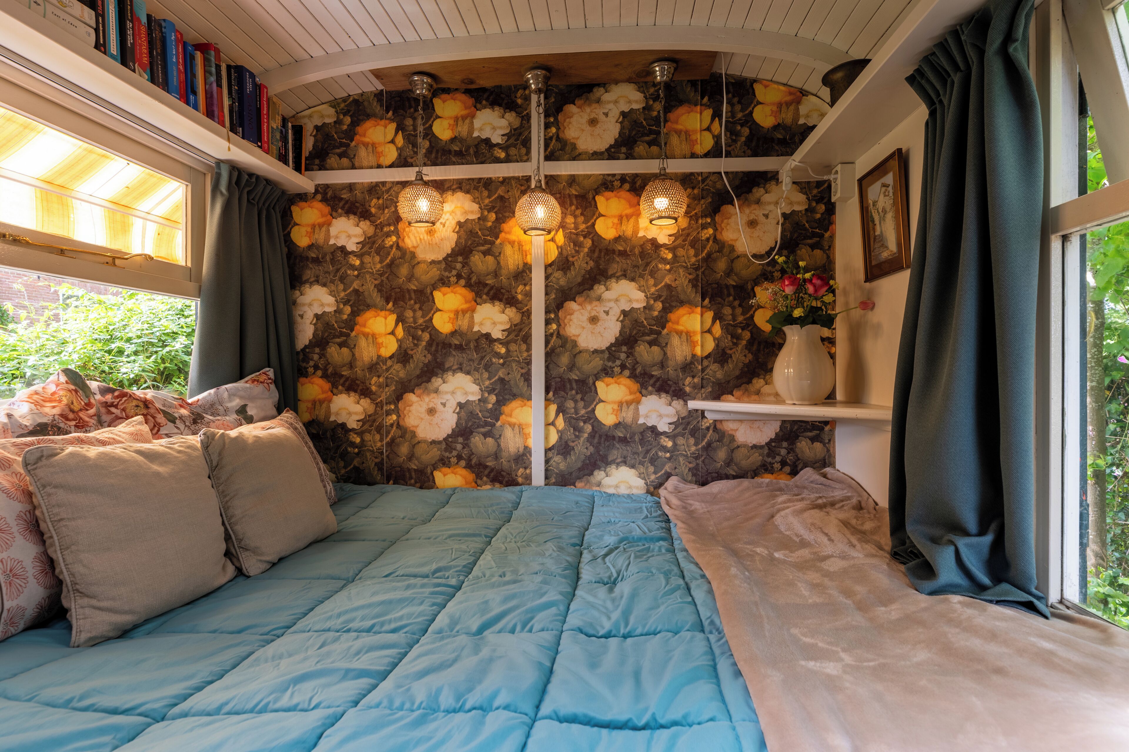 A bedroom with a blue bedspread, a stone wall with floral wallpaper, and a bookshelf with books.