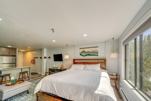 Ein modernes Hotelzimmer mit einem großen Bett, einem Schreibtisch mit Stuhl, einer kleinen Küchenzeile und einem großen Fenster mit Blick ins Freie.