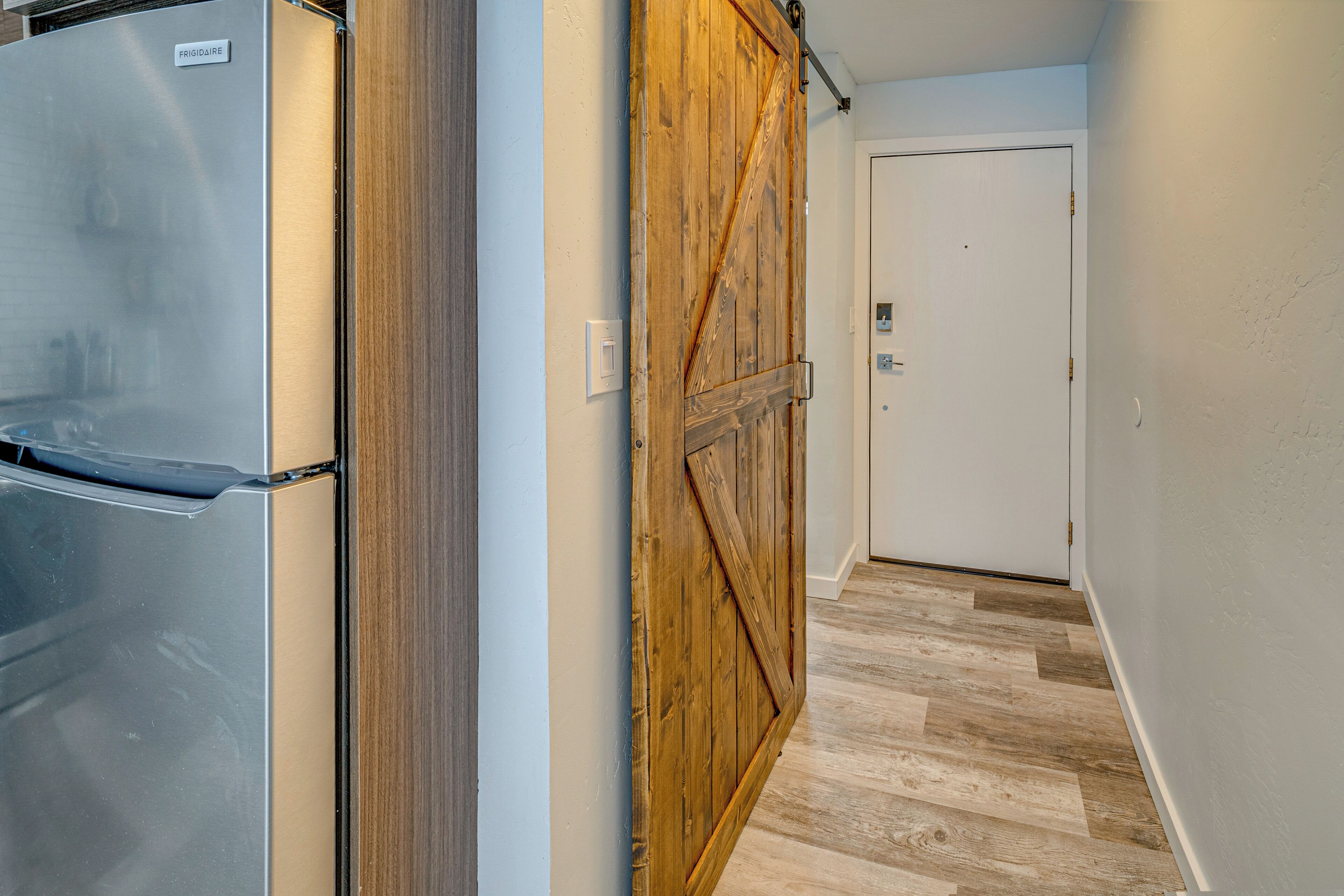 A hallway with a wooden door, a refrigerator, and a light switch on the wall.