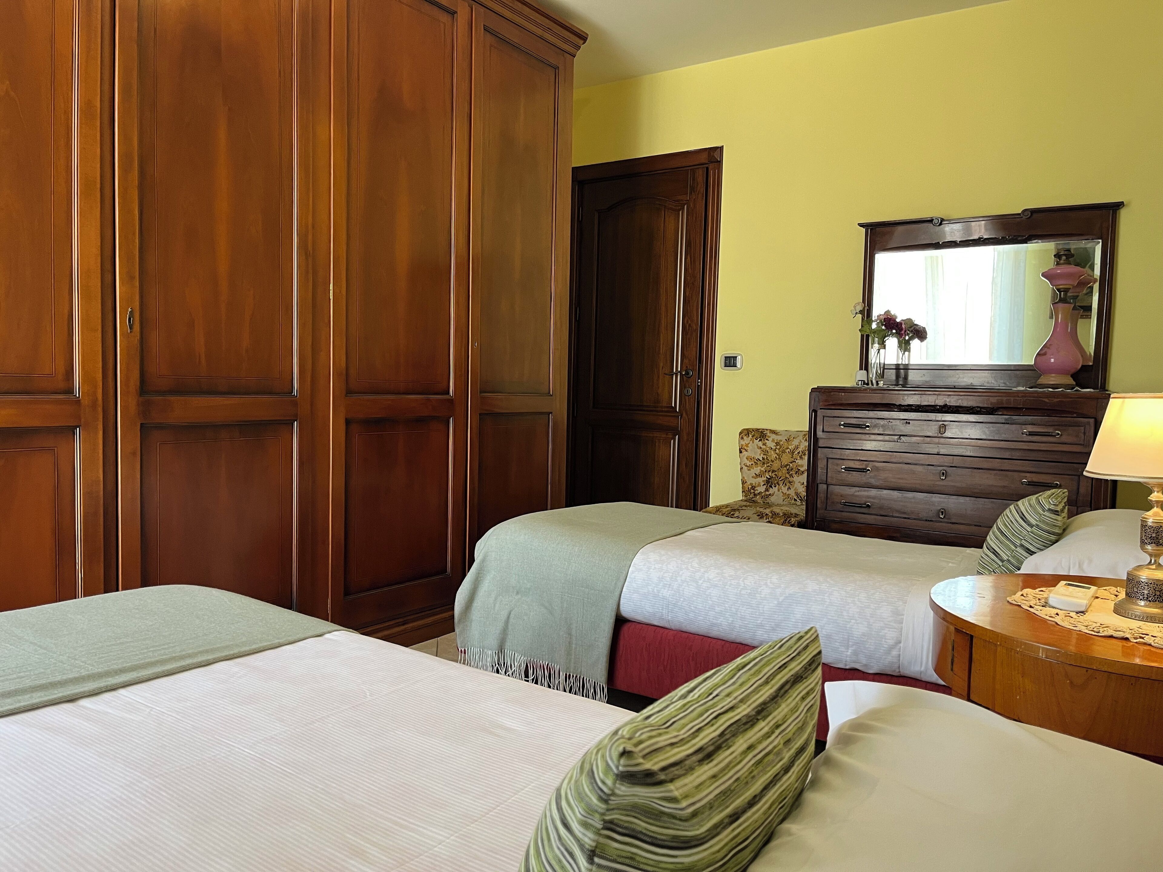 Quarto de hotel com duas camas de solteiro, um guarda-roupa de madeira, uma cômoda com espelho e uma mesa de cabeceira com abajur.
