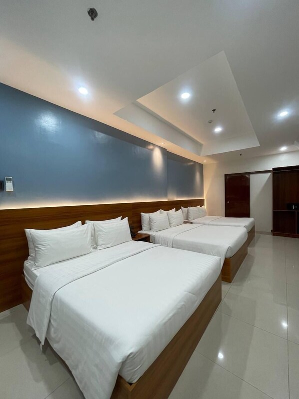 A hotel room with three beds, a wooden headboard, a large wall with a blue accent, and recessed ceiling lighting.