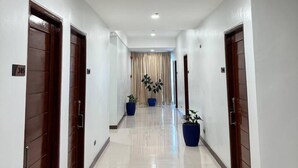 A long, well-lit hallway with white walls and wooden doors, leading to a room with a large potted plant.