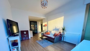 Een moderne woonkamer met een flatscreen televisie op een witte standaard, een grijze bank met oranje kussens, een salontafel en een potplant.
