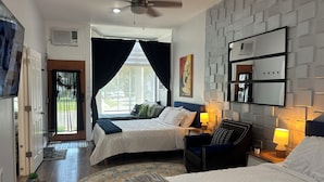 A hotel room with two beds, a ceiling fan, a large window with curtains, a wall-mounted TV, and a small side table with a lamp. - Historic Vintage House Downtown Grand Rapids  (Grand Rapids)