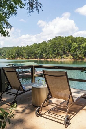 Une terrasse dotée de deux chaises longues et d’une petite table, offrant une vue sur un lac et une zone boisée.