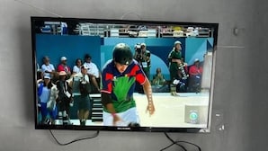 A falra szerelt televízión éppen egy sportesemény látható, alatta pedig egy polcon rágcsálnivalók találhatók.