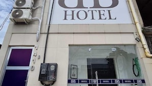 A szálloda homlokzatán feltűnő „BB HOTEL” felirat, légkondicionáló egységek és egy üvegportálos bejárat látható, melynek előterében egy virágtál áll.