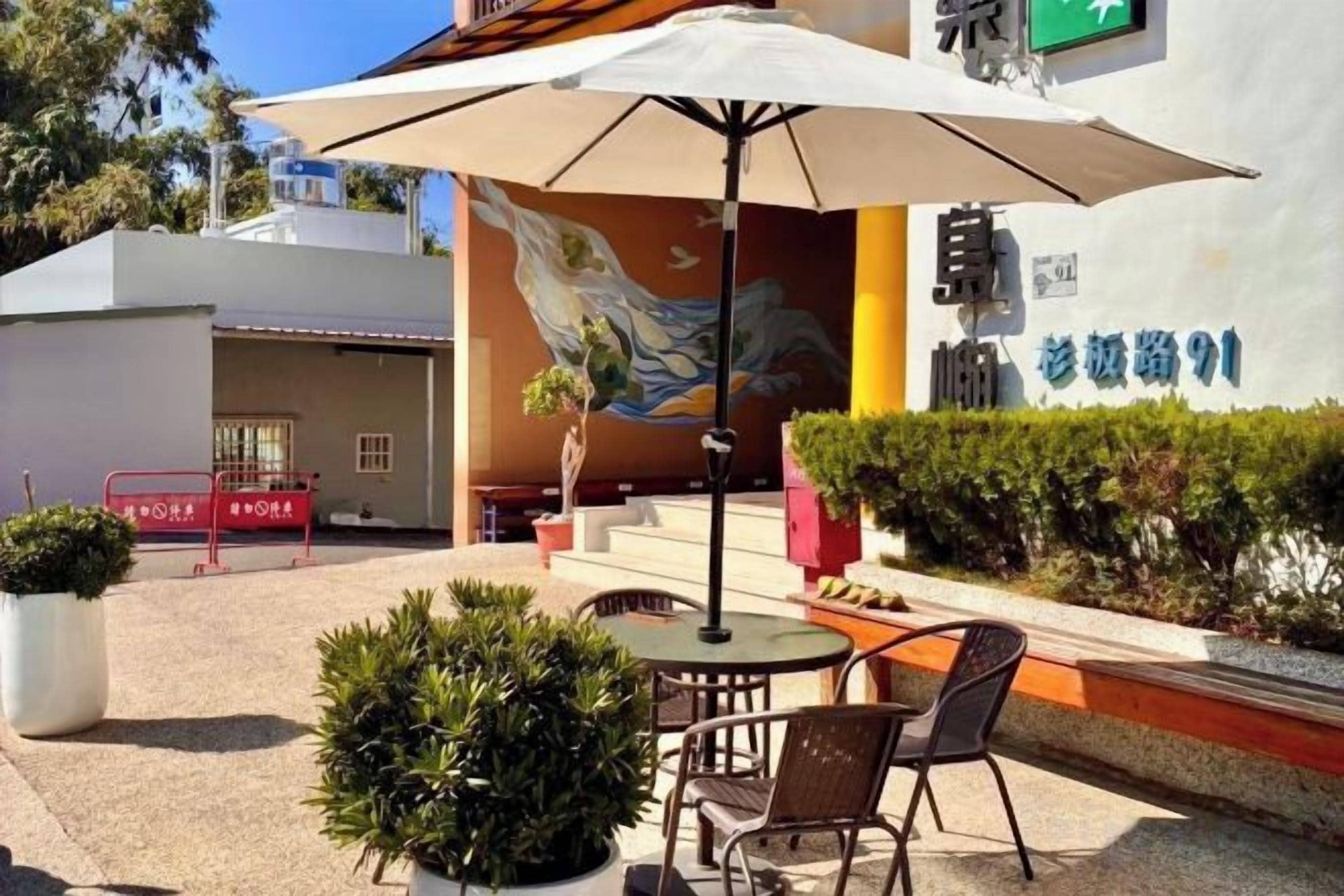 Un espace extérieur aménagé avec une table et des chaises sous un grand parasol, une fresque sur le bâtiment et une enseigne avec un numéro.