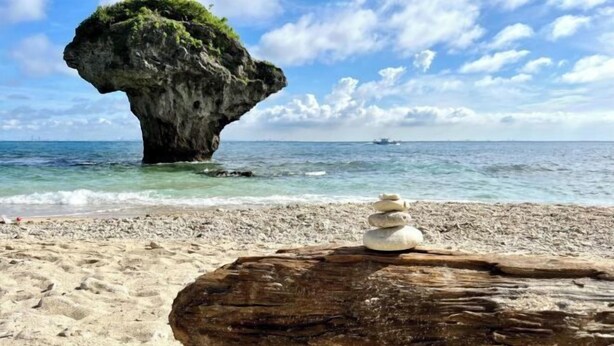 Una spiaggia con una grande formazione rocciosa, acqua cristallina e una pila di pietre su un tronco.