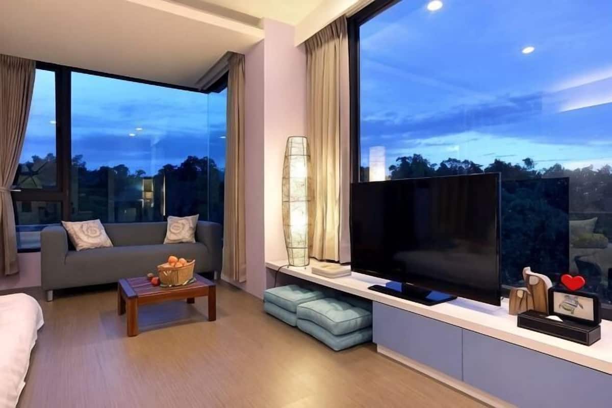 A modern living room with a sofa, a coffee table, a TV, and a large window offering a view of the outdoors.