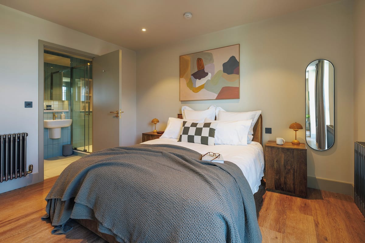 A modern bedroom with a large bed, a bedside table with a lamp, a mirror, and a bathroom visible through an open door.