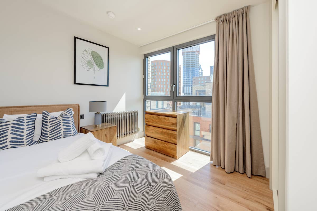 A modern hotel room with a large bed, a wooden dresser, a radiator, and a city view through a large window.