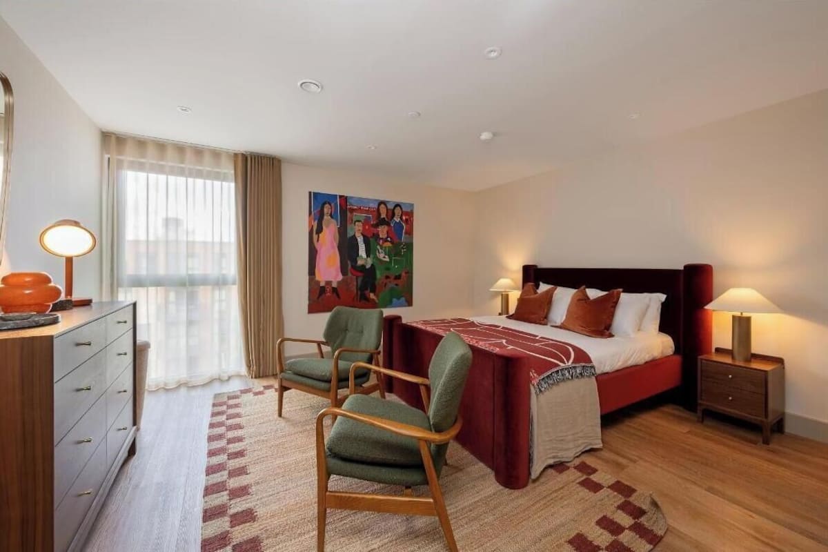 A hotel room with a bed, two chairs, a desk, a lamp, and a painting on the wall.
