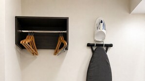 A room with a black ironing board, a shelf with hangers, and a wall-mounted ironing board.