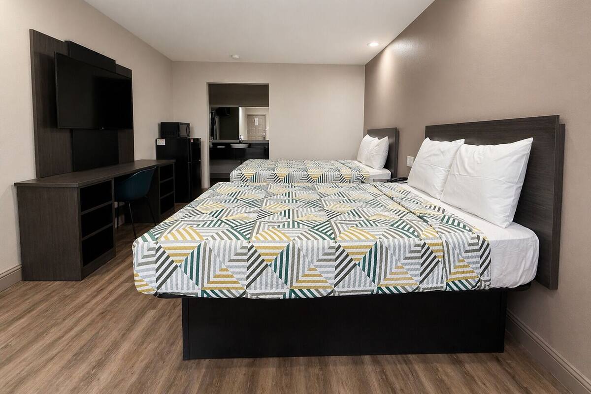 A hotel room with two beds, a TV, a desk, and a kitchenette.