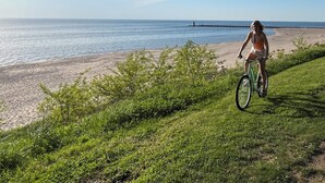 Une personne à vélo circule sur un sentier herbeux au bord d’une plage, d’où l’on a une vue dégagée sur l’océan et où l’on aperçoit une jetée au loin.