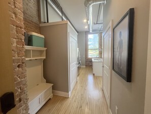 A narrow hallway with a wooden floor, white cabinetry, a brick wall, and a window with blinds.
