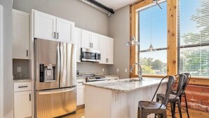Une cuisine moderne avec des meubles blancs, des appareils en acier inoxydable, un îlot central doté d’un comptoir en granit et une grande fenêtre avec des stores.