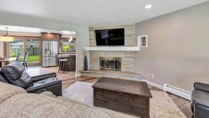 A living room with a stone fireplace, a flat-screen TV, a leather sofa, and a dining area visible through an open door. - Charming 4-Bedroom Home, Short Walk to Lambeau (Green Bay)