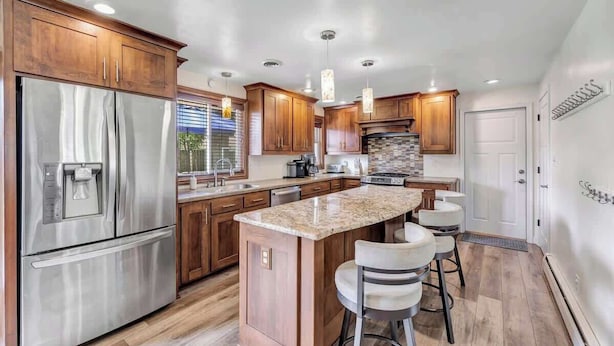 A modern kitchen with wooden cabinets, stainless steel appliances, and a central island with bar stools. - Charming 4-Bedroom Home, Short Walk to Lambeau (Green Bay)