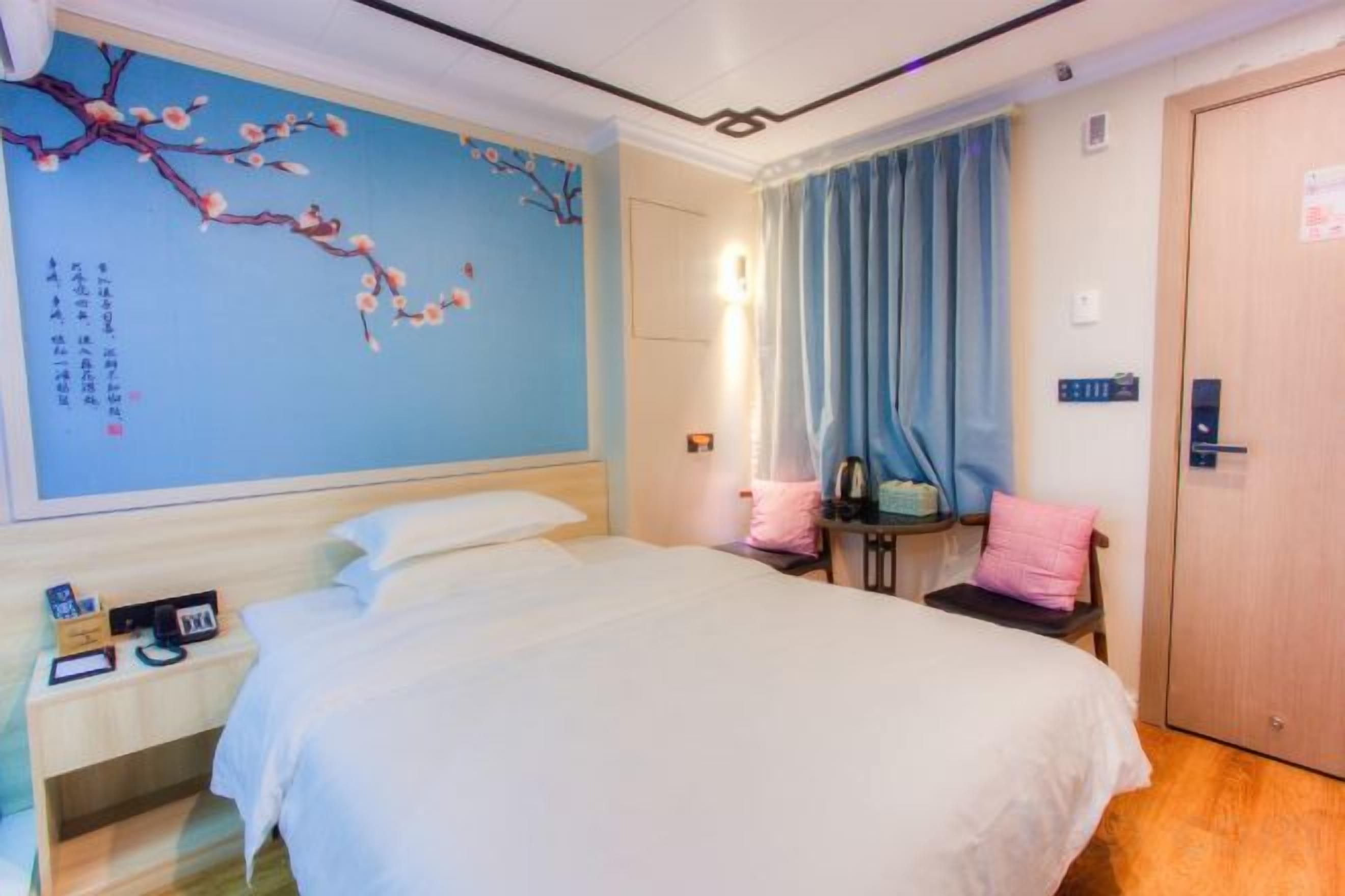 A hotel room with a large bed, a nightstand, a desk, a chair, a wardrobe, and a wall with a decorative painting.