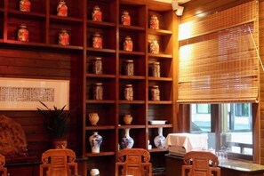 Una habitación con una estantería de madera llena de frascos de cerámica, un juego de té tradicional y una ventana con persianas de bambú.