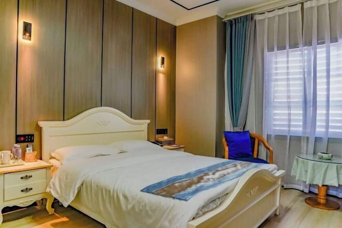 A hotel room with a bed, bedside tables, a chair, a small table, and a window with curtains.