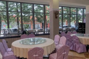 A dining area with round tables and chairs, large windows with a view of palm trees and parked cars. - Renhua Shaozhou Impression Boutique Cultural Hotel (Renhua)