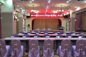 Un salón de conferencias con una larga mesa y sillas, un escenario con un cartel y una lámpara de araña.