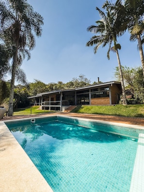 A swimming pool with clear blue water, surrounded by a tiled deck. In the background, there is a building with a flat roof and large windows. - Casa térrea com piscina e cozinha de cinema | Granja Viana (Jandira)