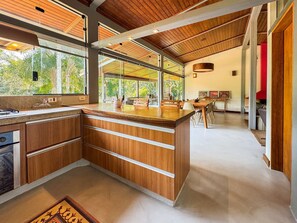 A modern kitchen with wooden cabinetry, a large island, and a view of the outdoors. - Casa térrea com piscina e cozinha de cinema | Granja Viana (Jandira)