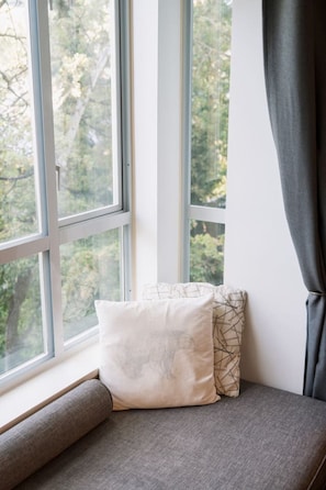 Un siège de fenêtre agrémenté d’un coussin et d’un oreiller à motifs, un rideau gris et une vue sur les arbres à l’extérieur.